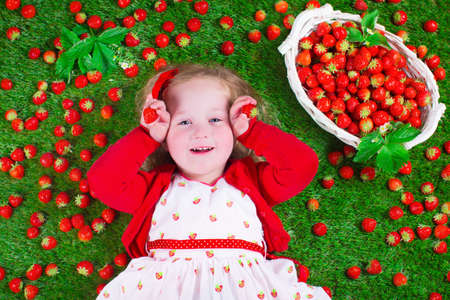 Child eating strawberry. Little girl playing peek a boo holding fresh ripe strawberries. Kids eating fruit relaxing on a lawn. Children summer fun on a farm picking berry.の写真素材