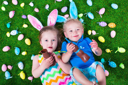 Children on Easter egg hunt. Kids eat chocolate rabbit. Boy and girl relaxing on a green lawn in the garden. Child enjoying sweets. Kid with bunny ears.の写真素材