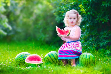 Child eating watermelon in the garden. Kids eat fruit outdoors. Healthy snack for children. Little girl playing in the garden holding a slice of water melon. Kid gardening.の写真素材