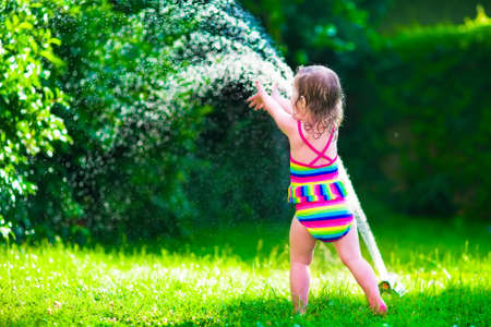 Child playing with garden sprinkler. Kid in bathing suit running and jumping. Kids gardening. Summer outdoor water fun. Children play with gardening hose watering flowers.の写真素材