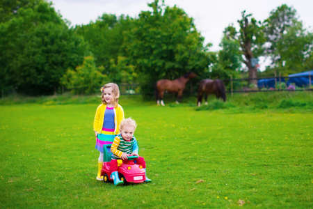 Family on a horse farm in summer. Brother and sister riding a toy car. Outdoor fun for children. Kids playing with pets. Child feeding an animal. Girl and little boy play together in the garden.の写真素材