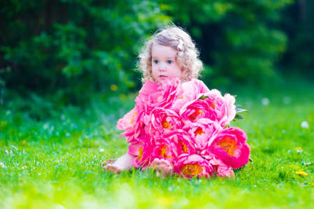 Little cute girl with peony flowers. Child wearing a pink dress playing in a summer garden. Kids gardening. Children play outdoors. Toddler kid with flower bouquet for birthday or mother's day.の写真素材
