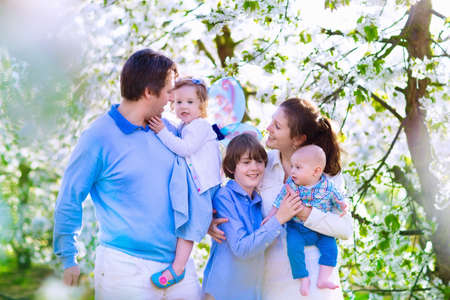 Family with kids in a blooming spring garden. Parents with three children baby toddler girl and boy enjoying picnic on a farm with apple and cherry trees. Mother father son and daughter in summerの写真素材