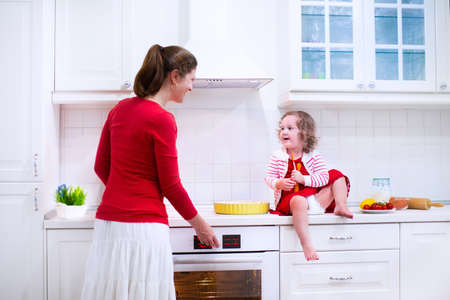 Mother and child bake a pie. Young woman and her daughter cook in a white kitchen. Kids baking pastry. Children helping to make dinner. Modern interior with oven and other appliances. Family eating.の写真素材