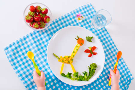 Healthy vegetarian lunch for little kids. Kid meal. Vegetable and fruit served as animals corn broccoli carrot strawberry helping child to learn eating right and clean children hands with spoonの写真素材