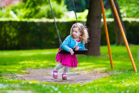 Little girl on a playground. Child playing outdoors in summer. Kids play on school yard. Happy kid in kindergarten or preschool. Children having fun at daycare play ground. Toddler on a swing.の写真素材