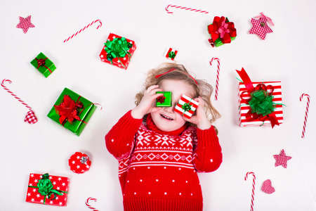 Child opening Christmas presents. Little girl in knitted winter sweater with present box. Kids open gifts. Toddler kid on the floor under decorated Xmas tree. Children play with gift box and candy.の写真素材