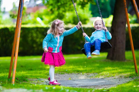 Little boy and girl on a playground. Child playing outdoors in summer. Kids play on school yard. Happy kid in kindergarten or preschool. Children having fun at daycare play ground. Toddler on a swing.の写真素材
