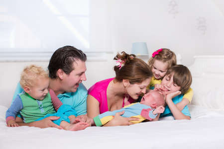 Big family with four kids relaxing in bed on a sunny morning. Parents with newborn baby, toddler boy, preschooler girl and teenager son in a white bedroom. Mother and father playing with children.の写真素材