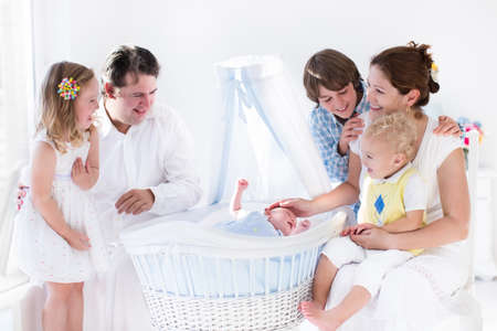 Big family with four kids in white bedroom. Parents and kids standing at crib of newborn baby boy....
