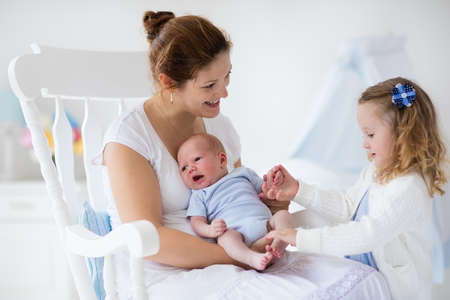 Little sister hugging her newborn brother. Toddler kid meeting new sibling. Mother and new born baby boy relax in a white bedroom. Family with children at home. Love, trust and tenderness concept.の写真素材