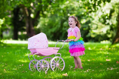 Little girl pushing toy stroller with bear. Toddler kid in pink dress playing with doll buggy. Kids birthday party. Children play outdoors. Mother and baby role game. Family summer fun. Preschool toysの写真素材
