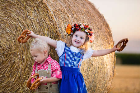 Kids in traditional Bavarian costumes in wheat field. German children eating bread and pretzel during Oktoberfest in Munich. Brother and sister play at hay bales during autumn harvest time in Germanyの写真素材