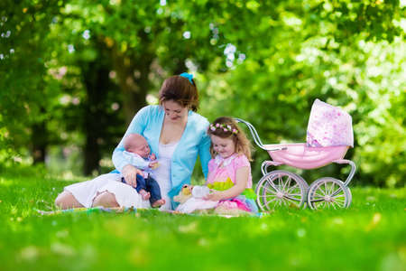 Family with children enjoying picnic outdoors. Mother with newborn baby and toddler child relax in a park. Little girl playing with toy stroller. Mom and kid play with infant boy. Kids birthday party.の写真素材