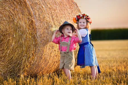 Kids in traditional Bavarian costumes in wheat field. German children eating bread and pretzel during Oktoberfest in Munich. Brother and sister play at hay bales during autumn harvest time in Germanyの写真素材