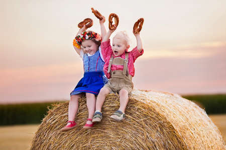 Kids in traditional Bavarian costumes in wheat field. German children eating bread and pretzel during Oktoberfest in Munich. Brother and sister play at hay bales during autumn harvest time in Germanyの写真素材