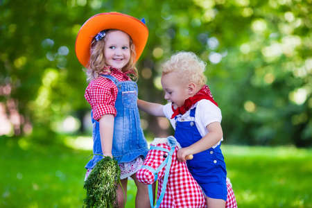 Little boy and girl dressed up as cowboy and cowgirl playing with toy rocking horse in park. Kids play outdoors. Children in Halloween costumes at trick or treat. Toys for preschooler or toddler childの写真素材