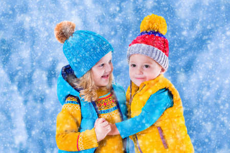 Little girl and boy in yellow and blue knitted hat catching snowflakes in winter park on Christmas eve. Kids play outdoor in snowy winter forest. Children catch snow flake on Xmas. Toddler kid playingの写真素材