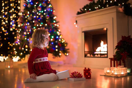 Family on Christmas eve at fireplace. Kids opening Xmas presents. Children under Christmas tree with gift boxes. Decorated living room with traditional fire place. Cozy warm winter evening at home.の写真素材