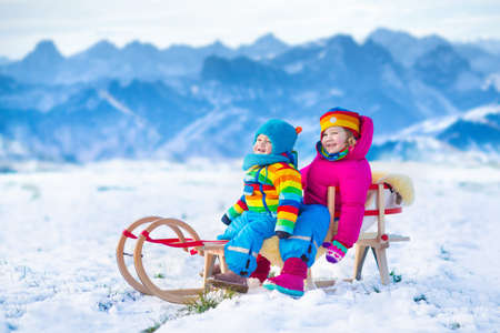 Little girl and baby boy enjoying a sleigh ride in the Alps mountains. Child sledding. Toddler kid riding a sledge. Children play outdoors in snow. Kids sled. Outdoor winter fun for Christmas vacationの写真素材