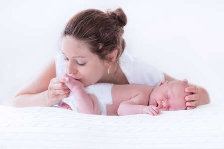 Young mother hugging her newborn child. Mom nursing baby. Woman and new born boy relax in a white bedroom. Family at home. Love, trust and tenderness concept. Bedding and textile for nursery.の写真素材