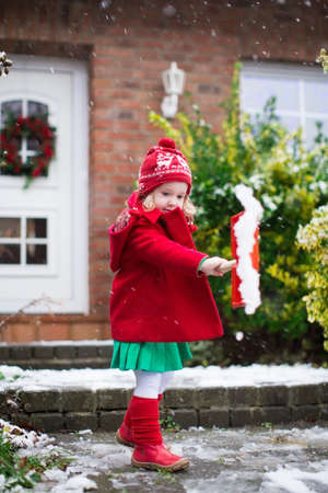 Little girl shoveling snow on home drive way. Beautiful house decorated for Christmas. Child with shovel playing outdoors in Xmas season. Family removing snow during blizzard. Kids play outside.の写真素材
