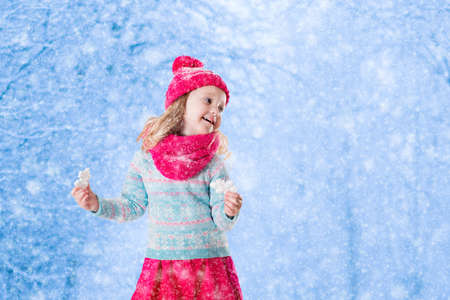 Little girl in blue knitted sweater and pink hat catching snowflakes in winter park. Kids play outdoor in snowy forest. Children catch snow flakes. Toddler kid playing outside in snow storm.の写真素材