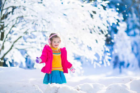 Funny little toddler girl in a colorful knitted hat and warm coat playing with snow. Kids play outdoors in winter. Children having fun at Christmas time. Child enjoying sunny day during Xmas vacation.の写真素材