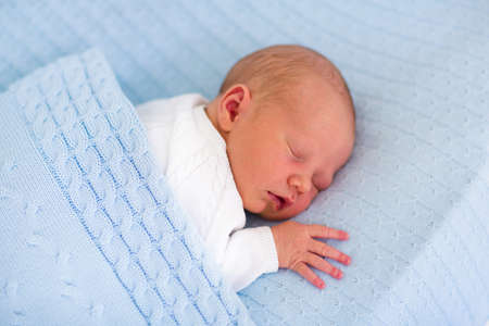 Newborn baby boy in bed. New born child sleeping under a blue knitted blanket. Children sleep. Bedding for kids. Infant napping in bed. Healthy little kid shortly after birth. Cable knit textile.の写真素材