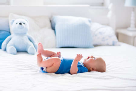 Adorable baby boy in white sunny bedroom. Newborn child relaxing in bed. Nursery for young children. Textile and bedding for kids. Family morning at home. New born kid during tummy time with toy bear.の写真素材