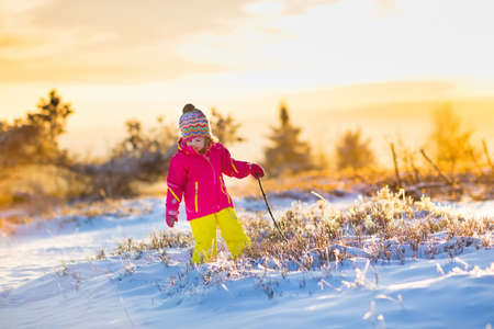 Child running in snowy forest. Toddler kid playing outdoors. Kids play in snow. Christmas vacation in sunny winter park for family with young children. Little girl in colorful jacket and knitted hat.の写真素材
