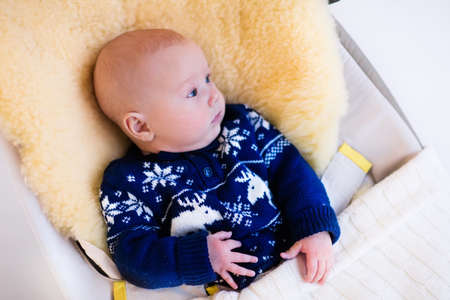 Funny little baby relaxing in a bouncer with sheepskin foot muff wearing warm knitted reindeer Christmas sweater at home on cold winter day. New born boy in stroller. Newborn child in rocking chair.の写真素材