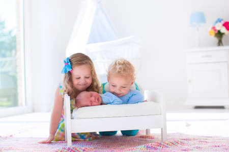 Cute little boy and girl kissing newborn brother. Toddler kids meet new born sibling at home. Infant sleeping in toy bed in white nursery. Kids playing and bonding. Children with small age difference.の写真素材