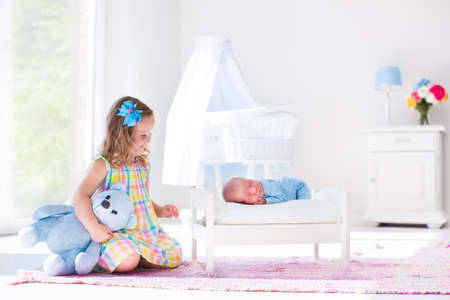 Cute little girl kissing newborn brother. Toddler kid meeting new born sibling. Infant sleeping in toy bed in white nursery. Kids playing. Siblings with small age difference. Children play and bond.の写真素材