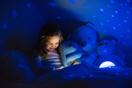 Little girl reading a book in bed. Dark bedroom with night light projecting stars on room ceiling....
