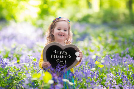 Child playing in bluebells forest. Little girl holding a wooden heart shape chalk board standing in a park with beautiful spring bluebell flowers. Copy space for your text. Kids having fun outdoors in spring.の写真素材