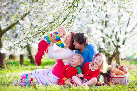 Big family with three little children eating lunch outdoors. Parents and kids with picnic basket in spring garden. Mother, father, preschooler girl, toddler boy and baby eat and drink in summer park.の写真素材