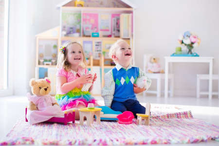 Kids playing with doll house and stuffed animal toys. Children sit on a pink rug in a play room at home or kindergarten. Toddler kid and baby with plush toy and dolls. Birthday party for little child.の写真素材
