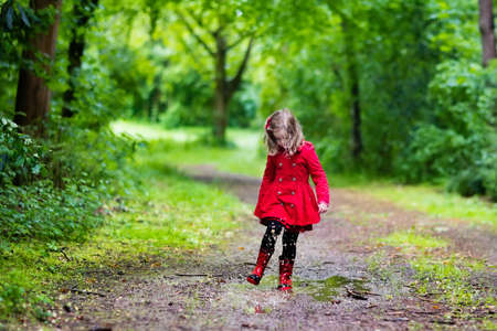 Little girl playing in rainy summer park. Child with red ladybug umbrella, waterproof coat and boots jumping in puddle and mud in the rain. Kid walking in autumn shower. Outdoor fun by any weather.の写真素材