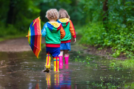 Little boy and girl play in rainy summer park. Children with colorful rainbow umbrella, waterproof boots jump in puddle and mud in the rain. Kids walk in autumn shower. Outdoor fun by any weatherの写真素材