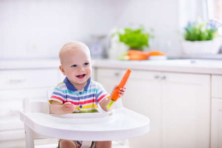 Happy baby sitting in high chair eating carrot in a white kitchen. Healthy nutrition for kids. Bio carrot as first solid food for infant. Children eat vegetables. Little boy biting raw vegetable.の写真素材