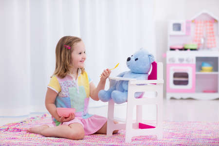 Little cute girl playing with her teddy bear. Child feeding doll in white nursery. Children play with wooden educational toys. Toddler kid in a playroom. Kids feed dolls and cook in toy kitchen.の写真素材