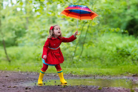 Little girl playing in rainy summer park. Child with colorful rainbow umbrella, waterproof coat and boots jumping in puddle in the rain. Kid walking in autumn shower. Outdoor fun by any weatherの写真素材