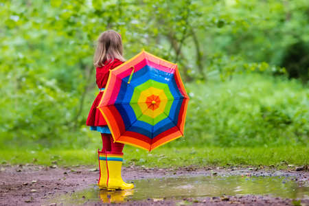 Little girl playing in rainy summer park. Child with colorful rainbow umbrella, waterproof coat and boots jumping in puddle in the rain. Kid walking in autumn shower. Outdoor fun by any weatherの写真素材
