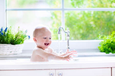 Baby taking bath in kitchen sink. Child playing with foam and soap bubbles in sunny bathroom with window. Little boy bathing. Water fun for kids. Hygiene and skin care for children. Bath room interiorの写真素材