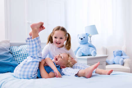 Happy kids playing in white bedroom. Little boy and girl, brother and sister play on the bed wearing pajamas. Nursery interior for children. Nightwear and bedding for baby and toddler. Family at home.の写真素材