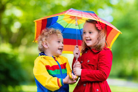 Little boy and girl play in rainy summer park. Children with colorful rainbow umbrella, waterproof jacket and coat playing in the rain. Kids walk in autumn shower. Outdoor fun by any weather.の写真素材