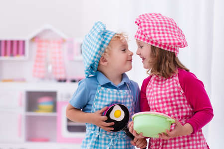 Little girl and boy in chef hat and apron cooking fried eggs in toy kitchen.  Wooden toys for young children. Kids play and cook at home or daycare. Toddler kid playing with stove, pans and dishes.の写真素材