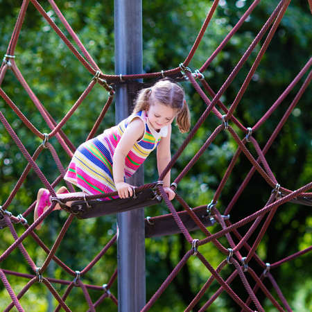 Active little child playing on climbing net and jumping on trampoline at school yard playground. Kids play and climb outdoors on sunny summer day. Cute girl on nest swing at preschool sport center.の写真素材