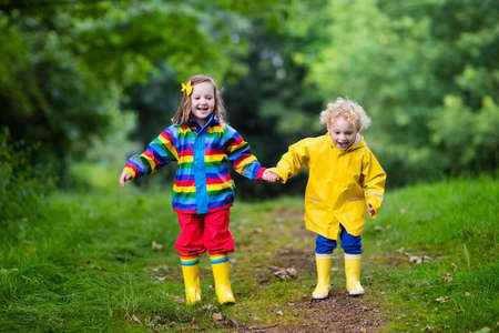 Little boy and girl play in rainy summer park. Children with colorful rainbow jacket and waterproof boots jump in puddle and mud in the rain. Kids walk in autumn shower. Outdoor fun by any weather.の写真素材